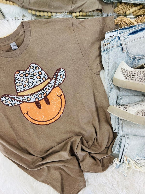 A brown short-sleeved graphic tee with a leopard wearing a cowboy hat and the text 'HAPPY' on it, displayed on a surface with jeans and shoes.