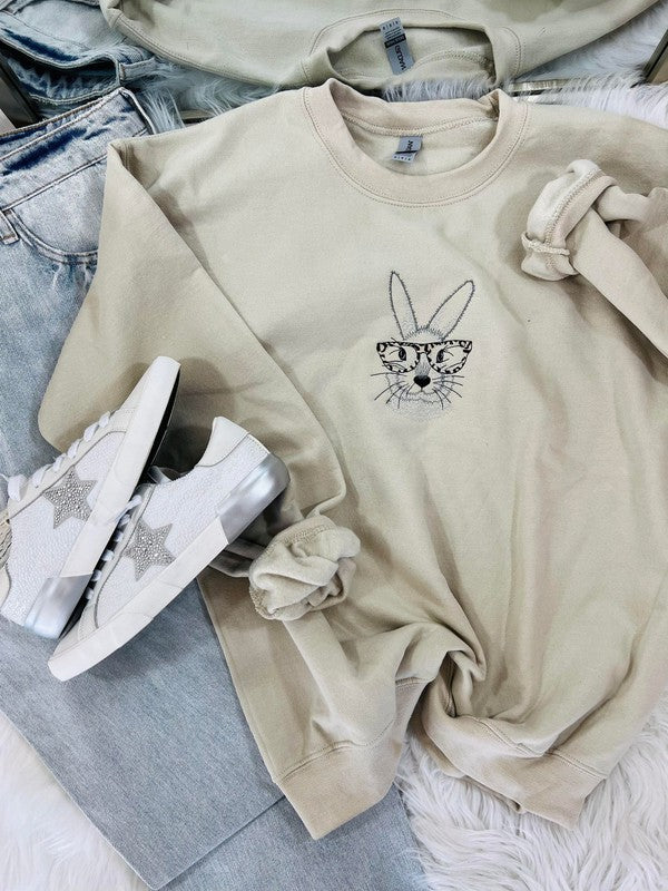A sand-colored embroidered sweatshirt with a graphic of a bunny wearing glasses, paired with white sneakers and blue jeans.