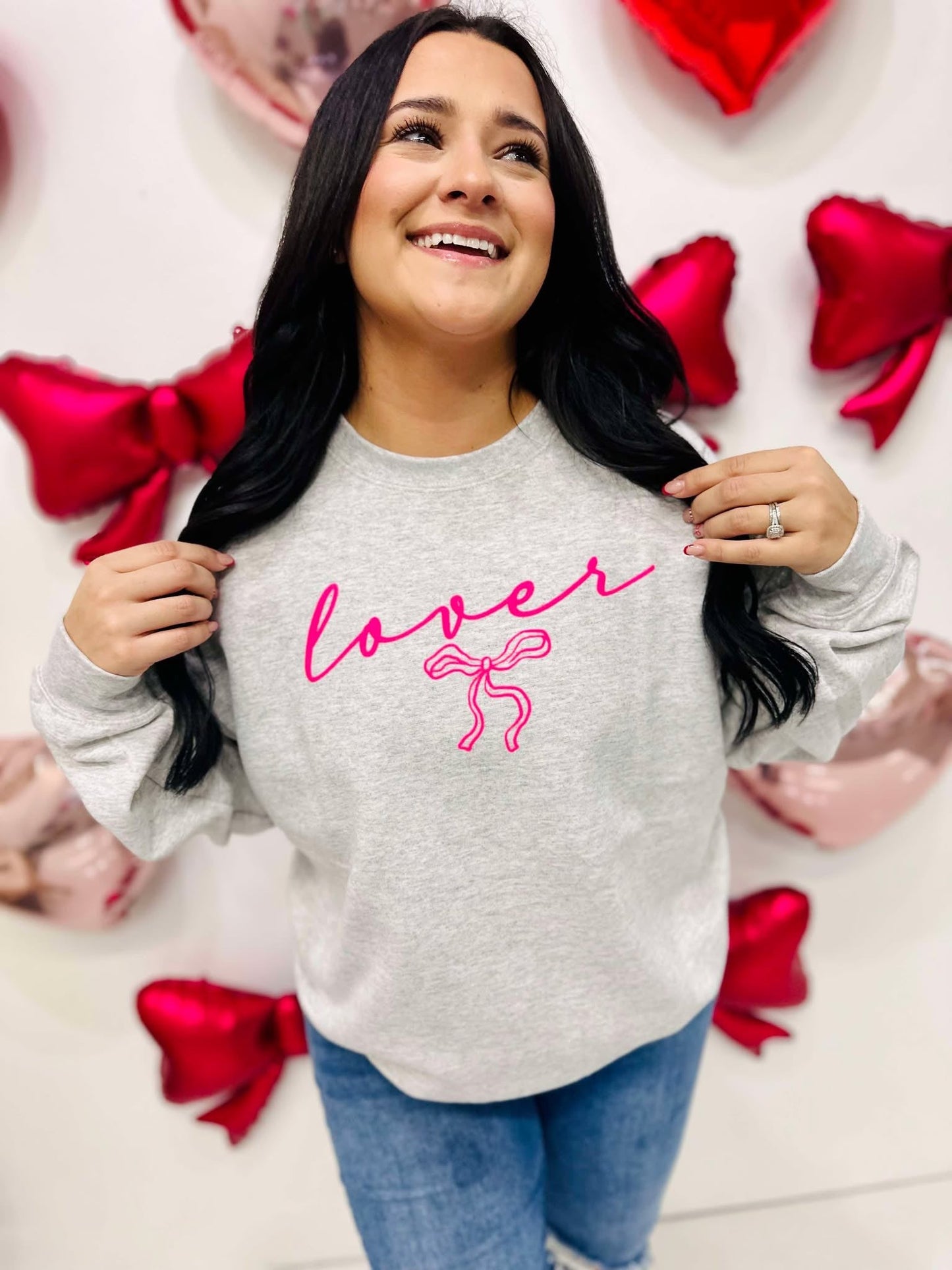 A woman smiling and posing with a gray sweatshirt that has the word 'lover' written in neon pink and a pink bow graphic.