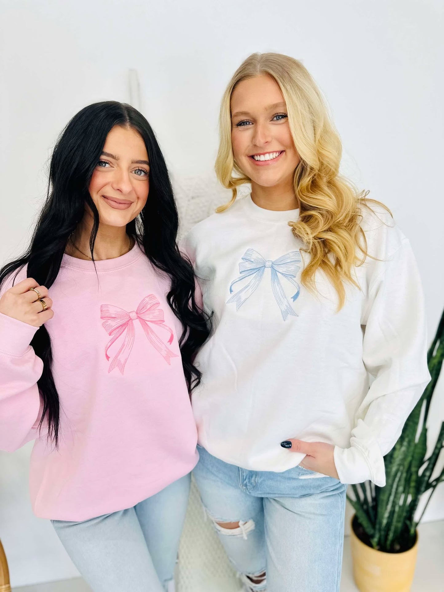 Two individuals standing next to each other, one wearing a pink and the other wearing a white embroidered bow sweatshirt.