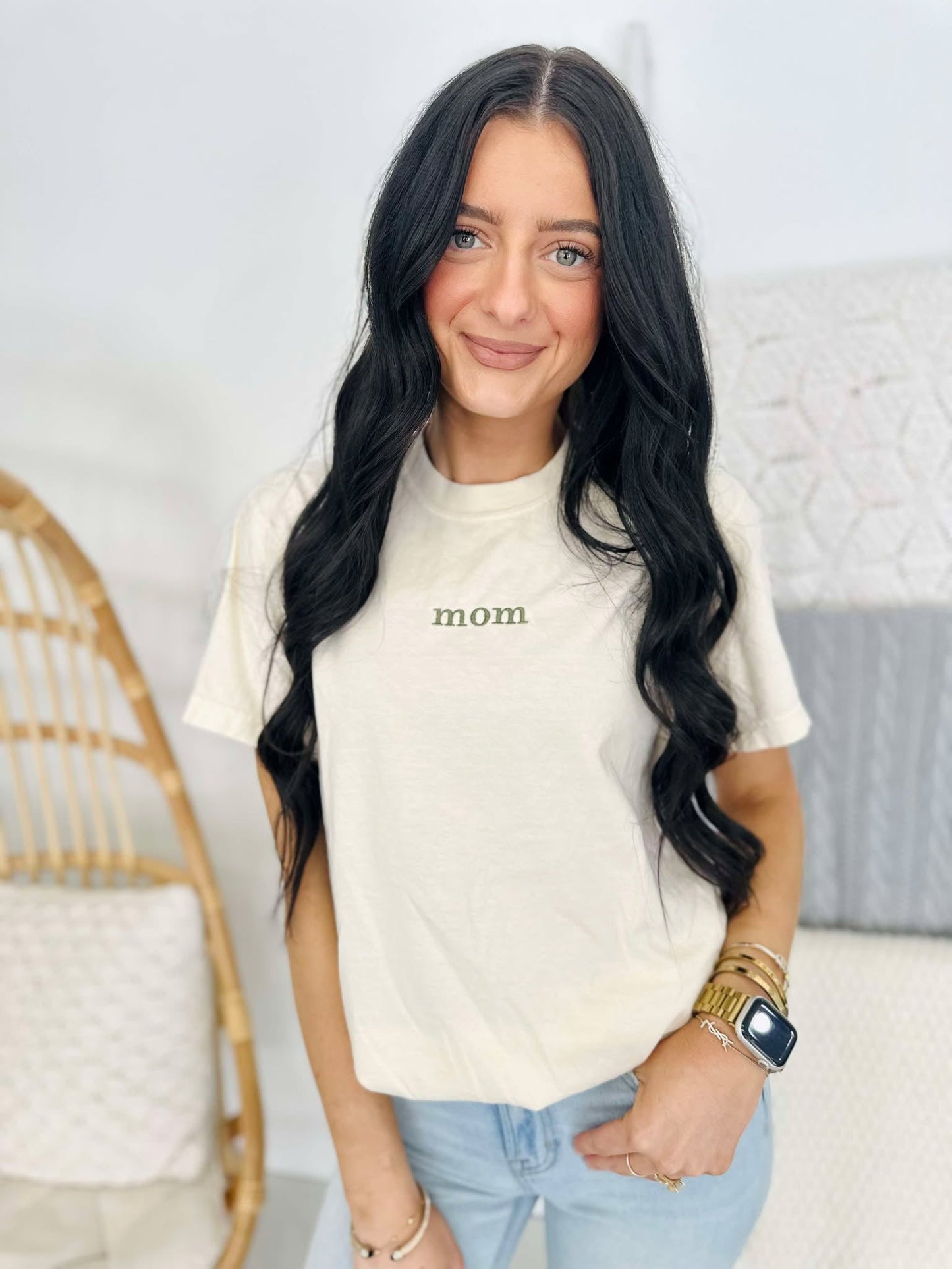 A woman wearing a white short-sleeved t-shirt with the word 'Mom' embroidered on the front, standing in a room with a wooden rocking chair and a watch on her wrist.