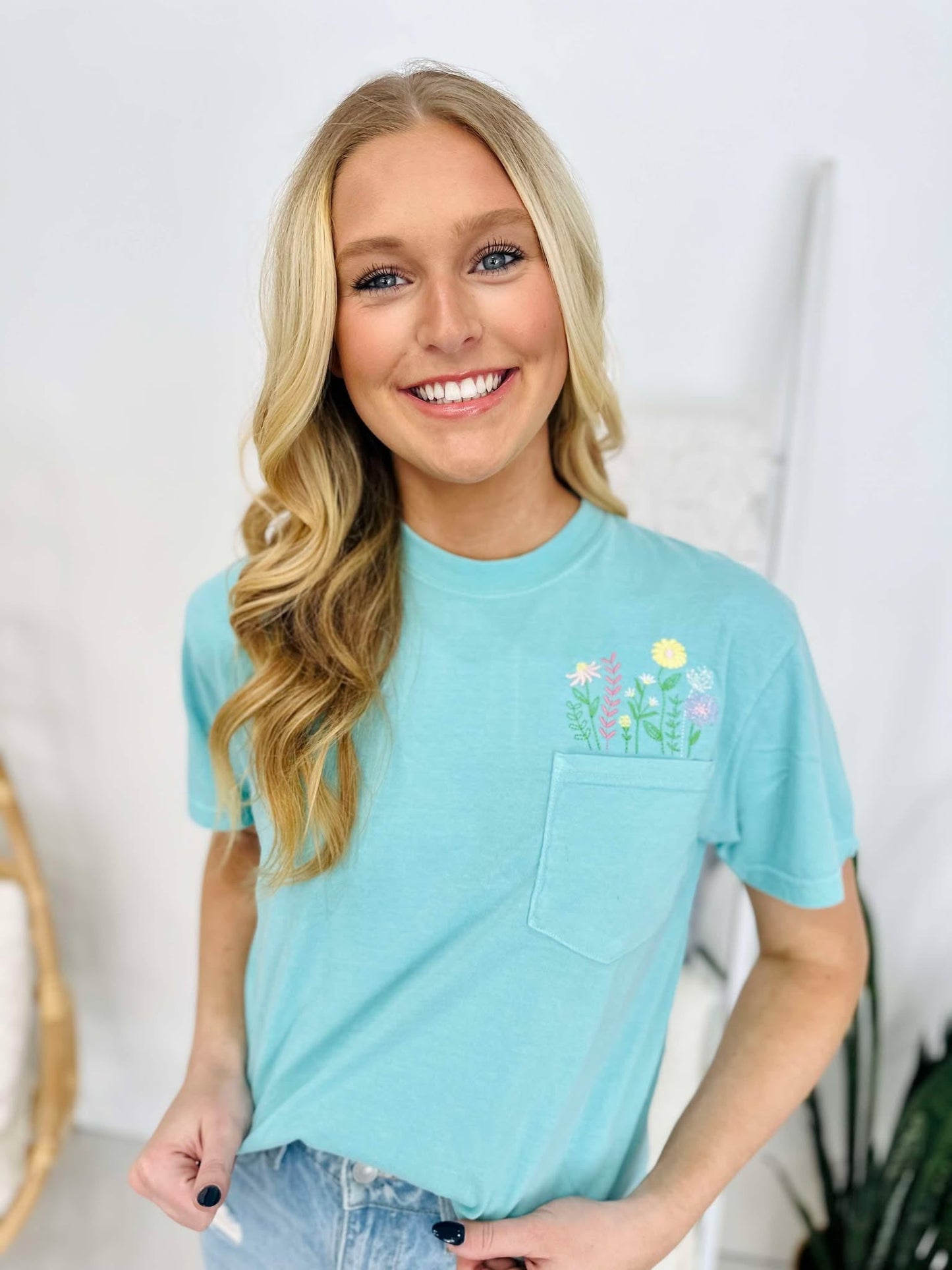 A woman wearing a chalky mint green t-shirt with a floral pocket design and embroidered lettering on the front, smiling against a white background.