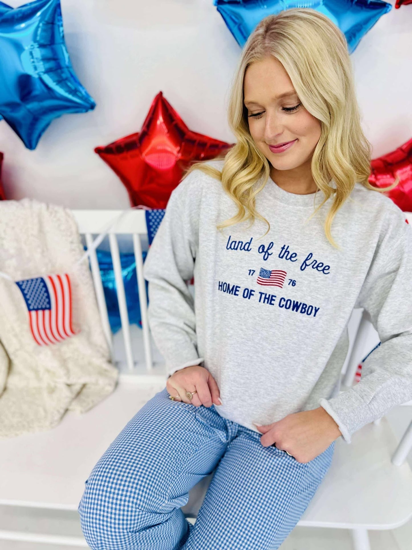 A woman sitting in front of a backdrop with American flag elements, wearing a gray sweatshirt with the text 'Land of the Free Home of the Cowboy' and stars.