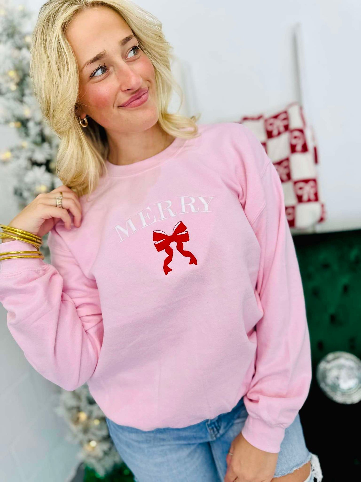 A woman wearing a light pink sweatshirt with 'Merry' embroidered on the front and a red bow detail.