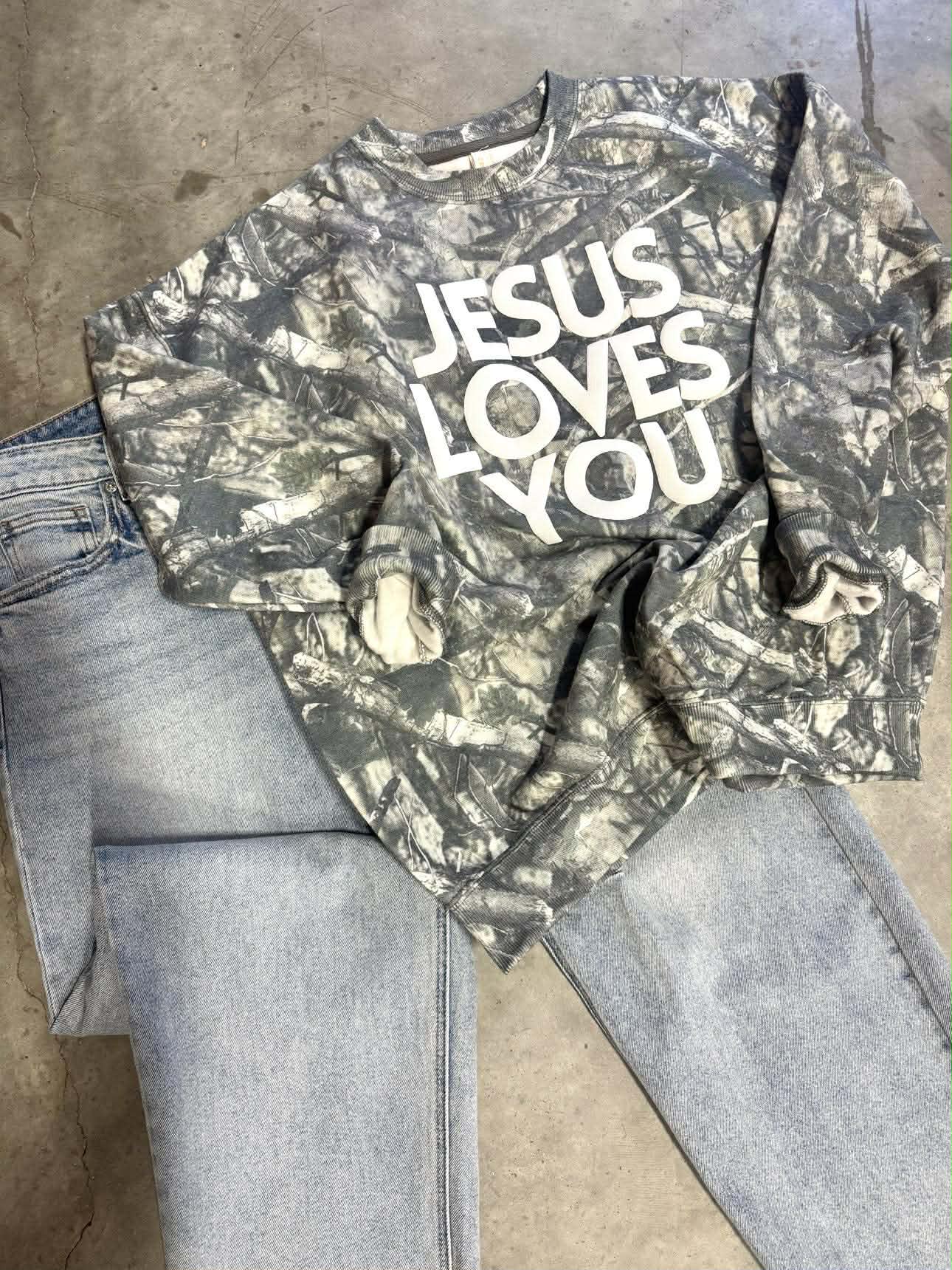 Camouflage long-sleeve shirt with 'Jesus Loves You' text on a concrete floor.