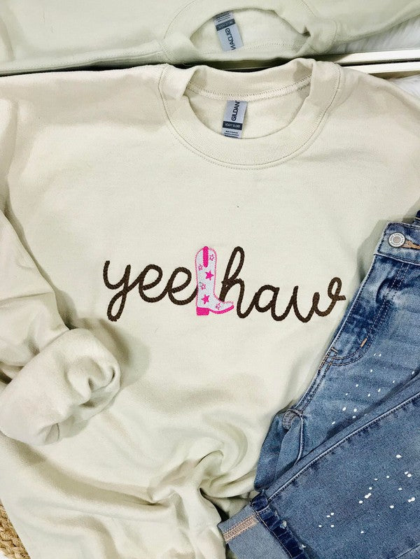 A tan embroidered sweatshirt with the text 'YEE HAW' on it, displayed on a surface with blue jeans and other clothing items.