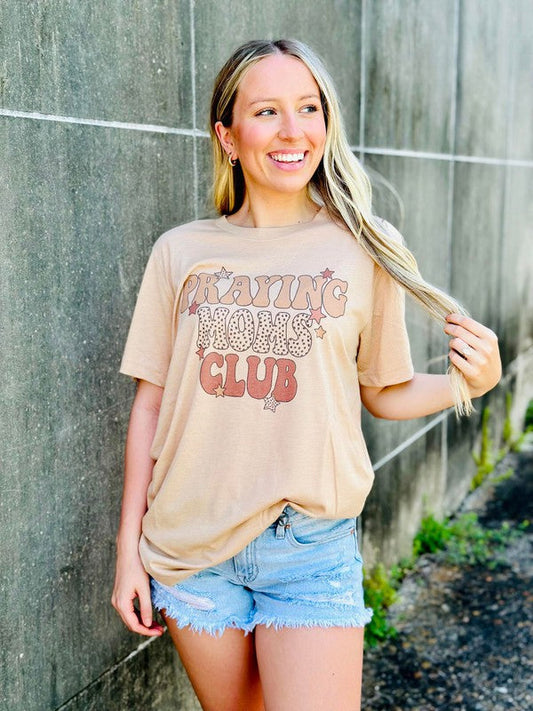 Woman wearing a beige t-shirt with text and denim shorts standing against a concrete wall Praying moms club.
