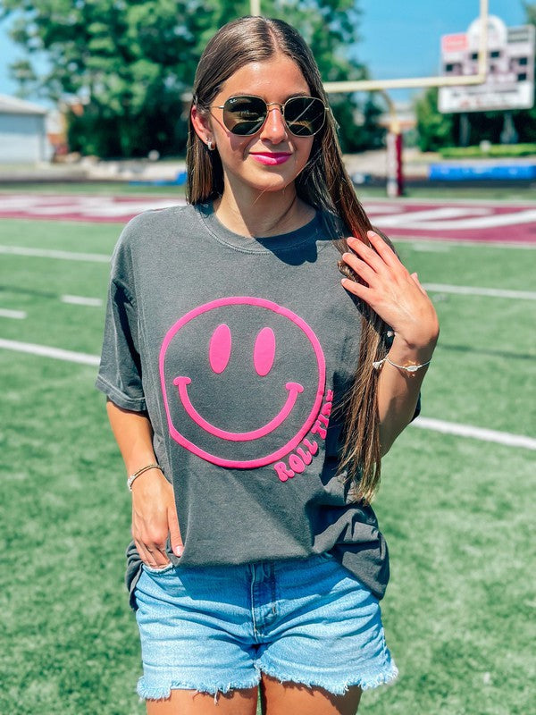 A female model wearing a gray t-shirt with a neon pink smiley face graphic on it, paired with denim shorts and mascot name, standing on an athletic field.