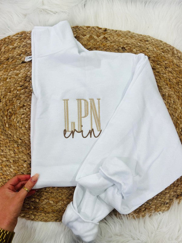 A white sweatshirt with the embroidered initial 'LPN' in brown on the chest area, displayed on a furry surface.
