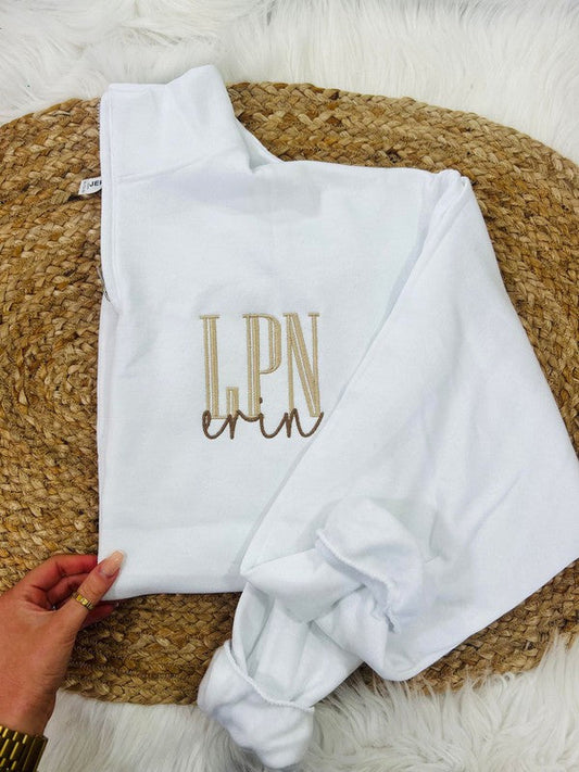 A white sweatshirt with the embroidered initial 'LPN' in brown on the chest area, displayed on a furry surface.