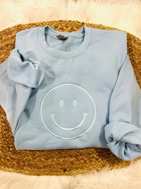 A light blue sweatshirt with an embroidered lettering of a smiling face on the chest, displayed on a woven mattress.
