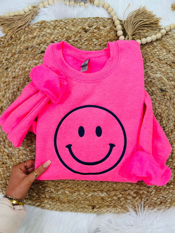 A neon pink sweatshirt with a black smiley face embroidered on the front, displayed on a furry surface.
