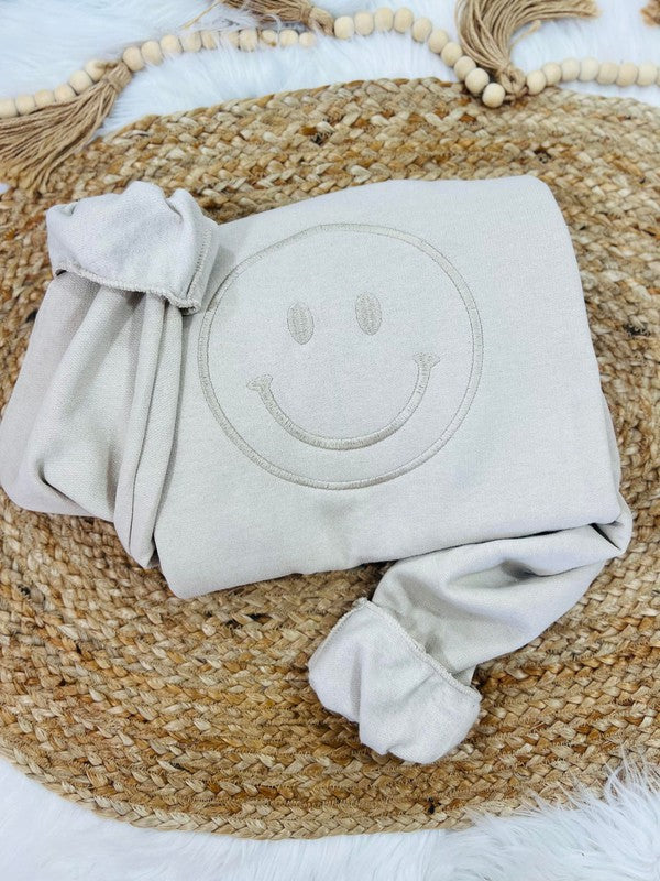 A beige embroidered sweatshirt with a happy face design on the front, displayed on a woven mat.