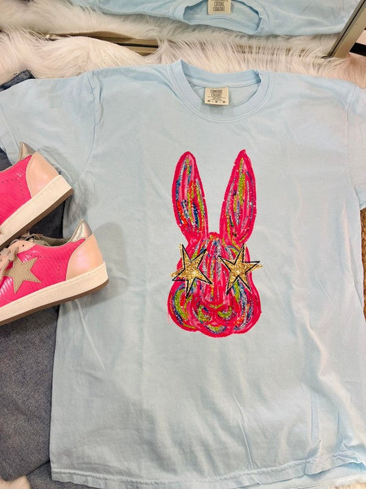 A light blue colored t-shirt with a graphic print of an eye-catching, colorful Easter bunny on it, displayed flat with a pair of pink sneakers and a white shoe on top of it.