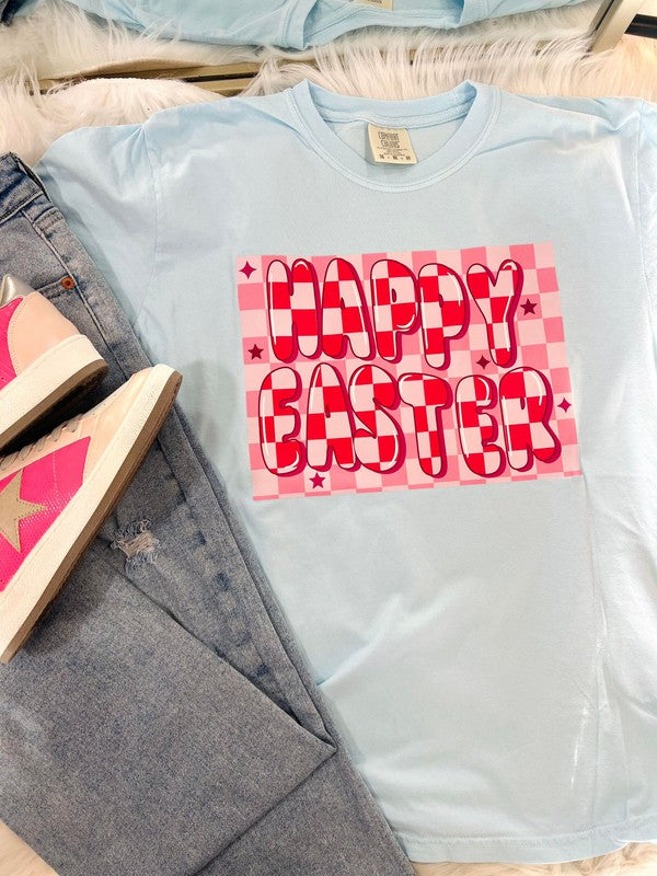A light blue colored t-shirt with a 'Happy Easter' text and pink checkered pattern on it, displayed on a flat surface with denim jeans and a wooden object in the background.