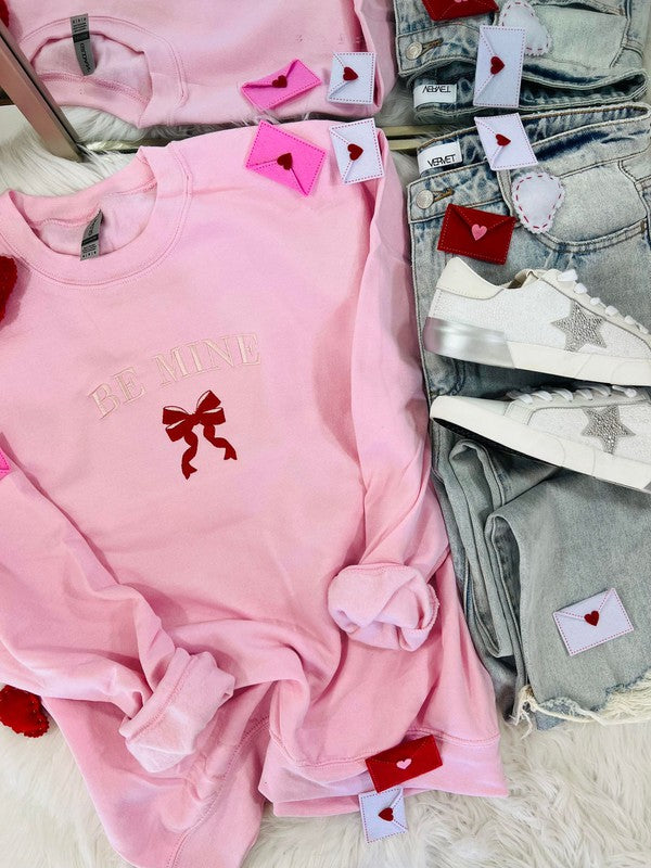 The image displays a light pink embroidered sweatshirt with the phrase "BE MINE" and a red ribbon design, presented next to a pair of blue denim jeans. The sweatshirt features decorative red heart-shaped tags and embroidered stars, enhancing its Valentine's Day-themed aesthetic.