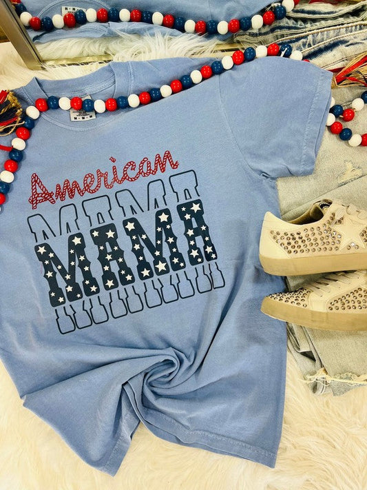 A blue t-shirt with the text 'American MAMA' printed on it, presented on a flat surface with decorative items around it.