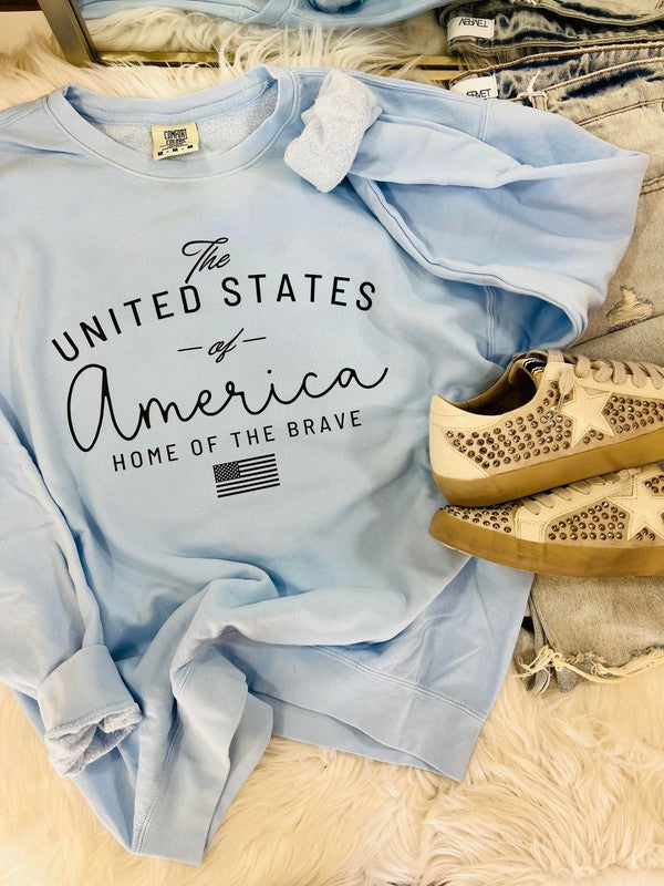 A light blue t-shirt with the text 'The United States of America Home of the Brave' printed on it, displayed flat with a pair of beige shoes at the bottom of the frame.