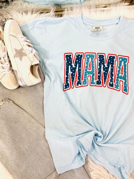 A blue curved-neck t-shirt with the word 'MAMA' and a star graphic in red and blue colors, displayed flat with a pair of white sneakers and a furry white blanket in the background.