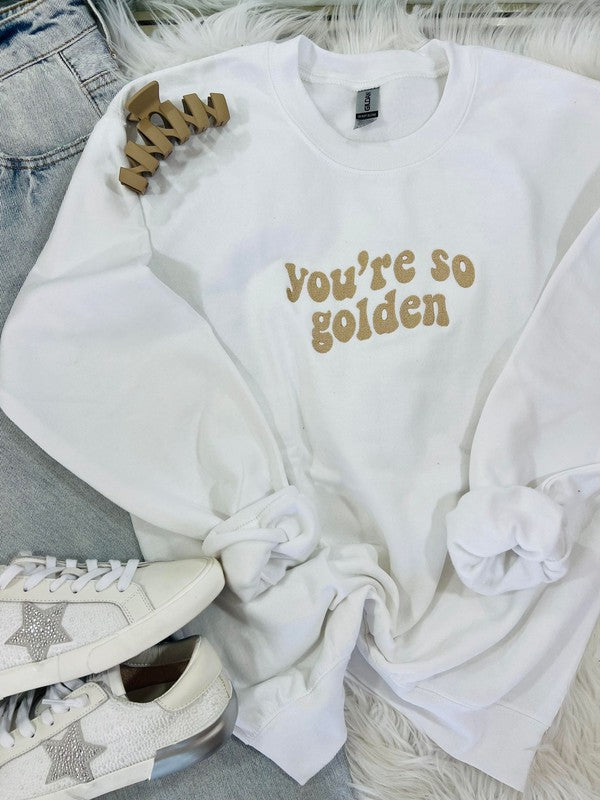 A white retro wave embroidered sweatshirt with the text 'you're so golden' on the front, displayed on a furry surface with a pair of sneakers and a star-shaped brooch.