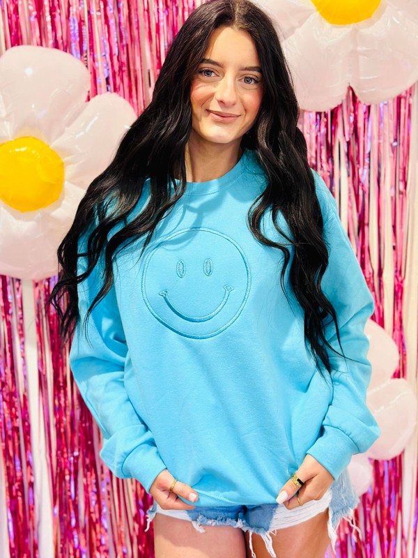 A person wearing a blue sweatshirt with a happy face embroidery on the front, posing against a pink glittery background with balloons.