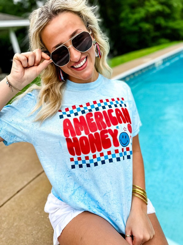 A person sitting by a poolside wearing a light blue graphic t-shirt with the text 'AMERICAN HONEY' in a patriotic design.