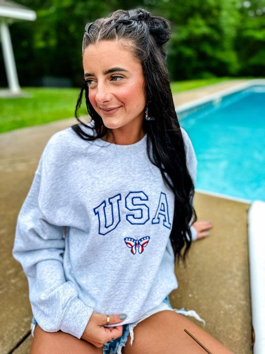 A person sitting by a poolside wearing an ash grey sweatshirt with the embroidered letters 'USA' and a butterfly logo on the front.