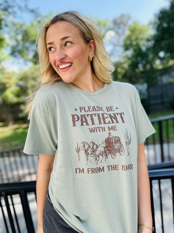 A woman wearing a green short-sleeved graphic tee with the text 'Please be patient with me I'm from the 1900's' printed on it, standing outdoors.