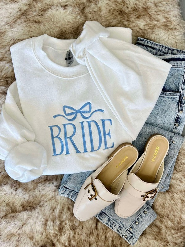 A white hoodie with the word 'Bride' embroidered on the front, paired with blue jeans and beige flats on a faux furry surface.