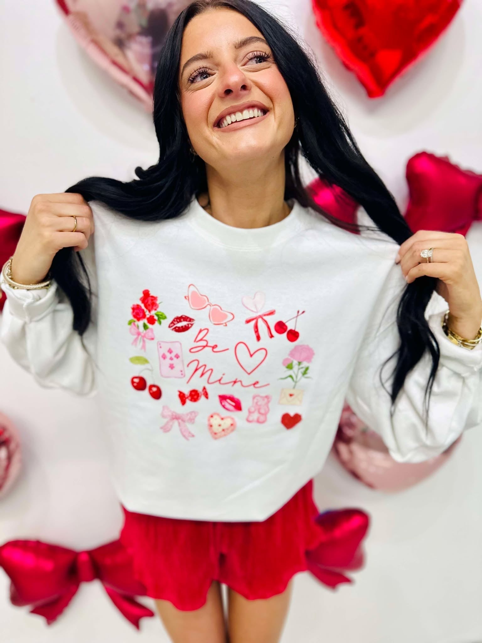 Woman wearing a white sweatshirt with Valentine's Day designs surrounded by heart-shaped decorations.