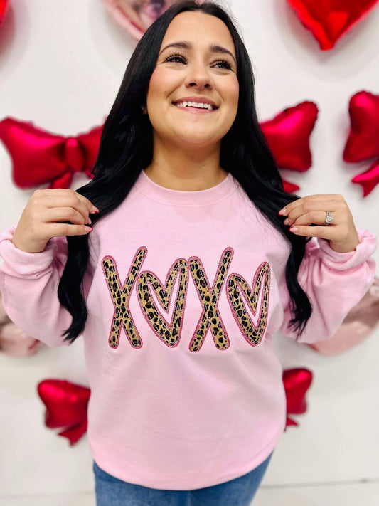 A person wearing a light pink sweatshirt with the word 'XOXO' embroidered in a leopard print style on the front, surrounded by heart-shaped decorations.