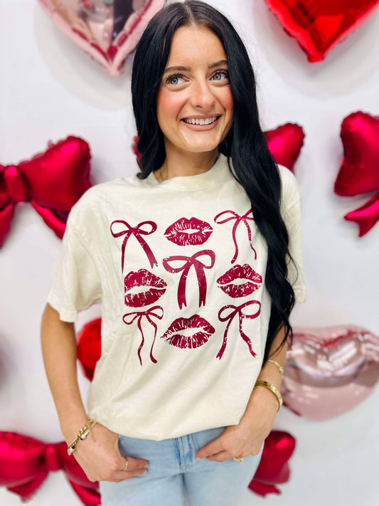 A person wearing a beige t-shirt with a graphic print of three stylized lips and bows in red and white colors, standing against a backdrop of Valentine's Day-themed decorations including balloons and hearts.