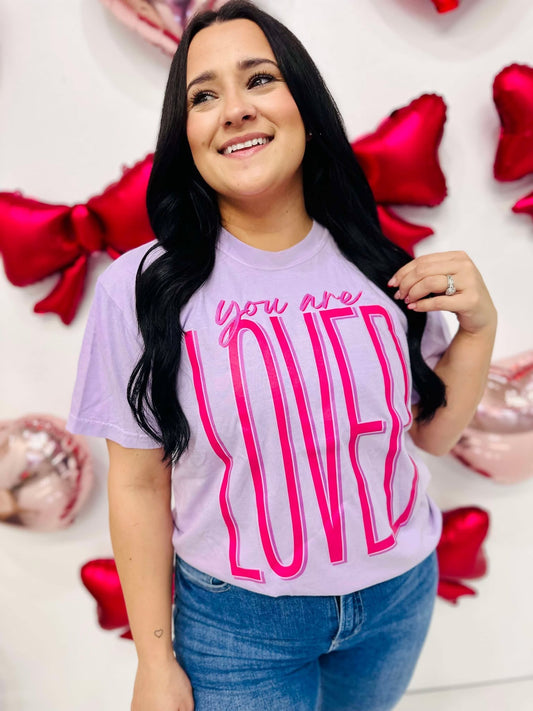 A woman smiling and wearing a purple t-shirt with the text 'you are LOVED' printed in large pink letters.