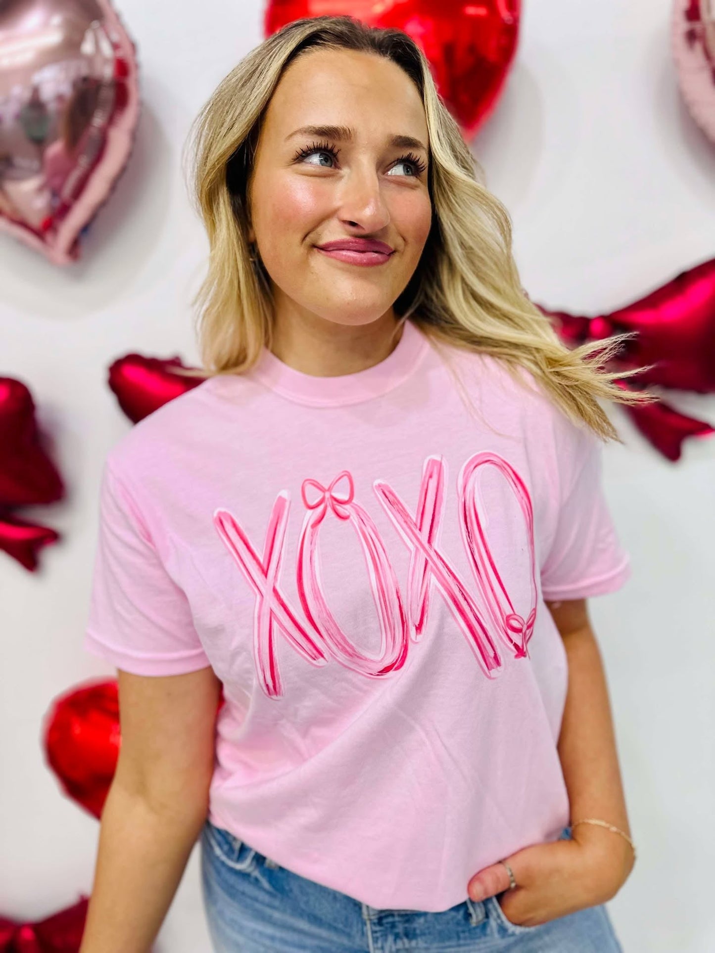 A woman wearing a pink t-shirt with the text 'XOXO' and a pink bow design on it, standing in a room with balloons and decorative hearts.
