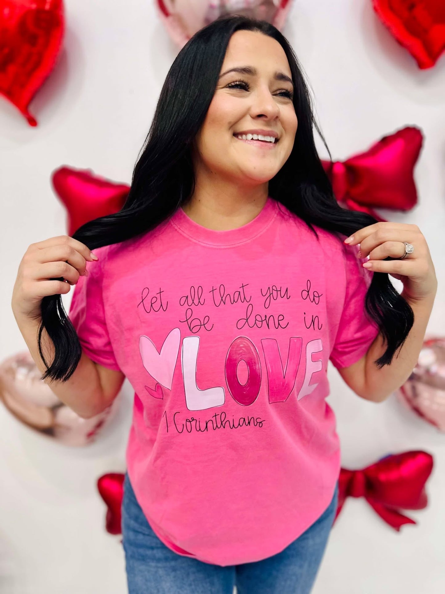 A person wearing a pink t-shirt with the text 'Let all you do be done in LOVE' printed on it, standing in a room with balloons and decorative hearts.