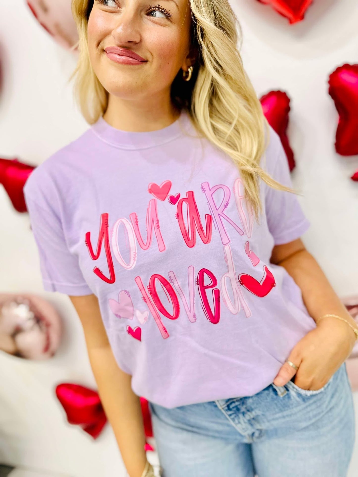 Woman wearing a purple t-shirt with 'You are love' text and hearts, surrounded by red heart-shaped props.
