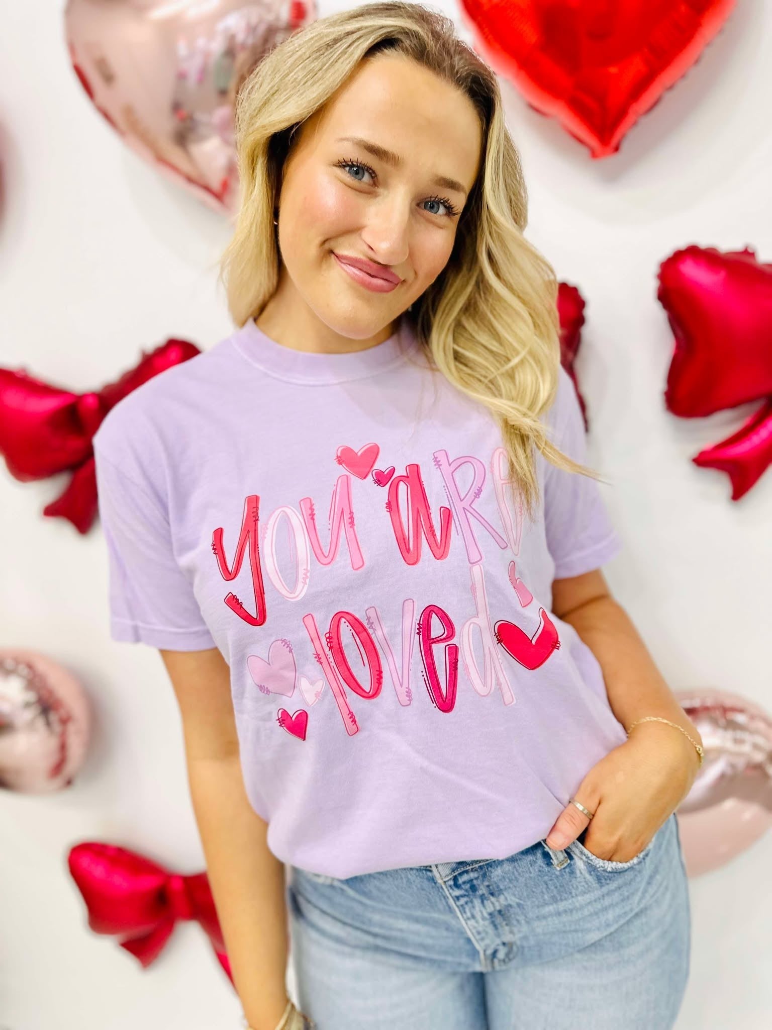 A person wearing a purple graphic tee with the text 'you're loved' and heart doodles, posing with a background of red and white heart-shaped decorations.