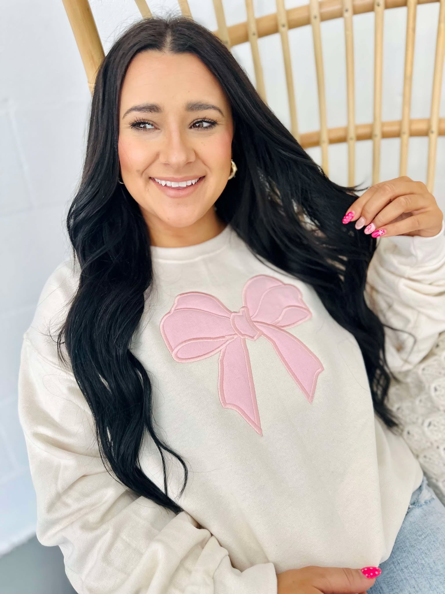 A woman smiling and posing in a beige sweatshirt with a pink bow applique on the front.