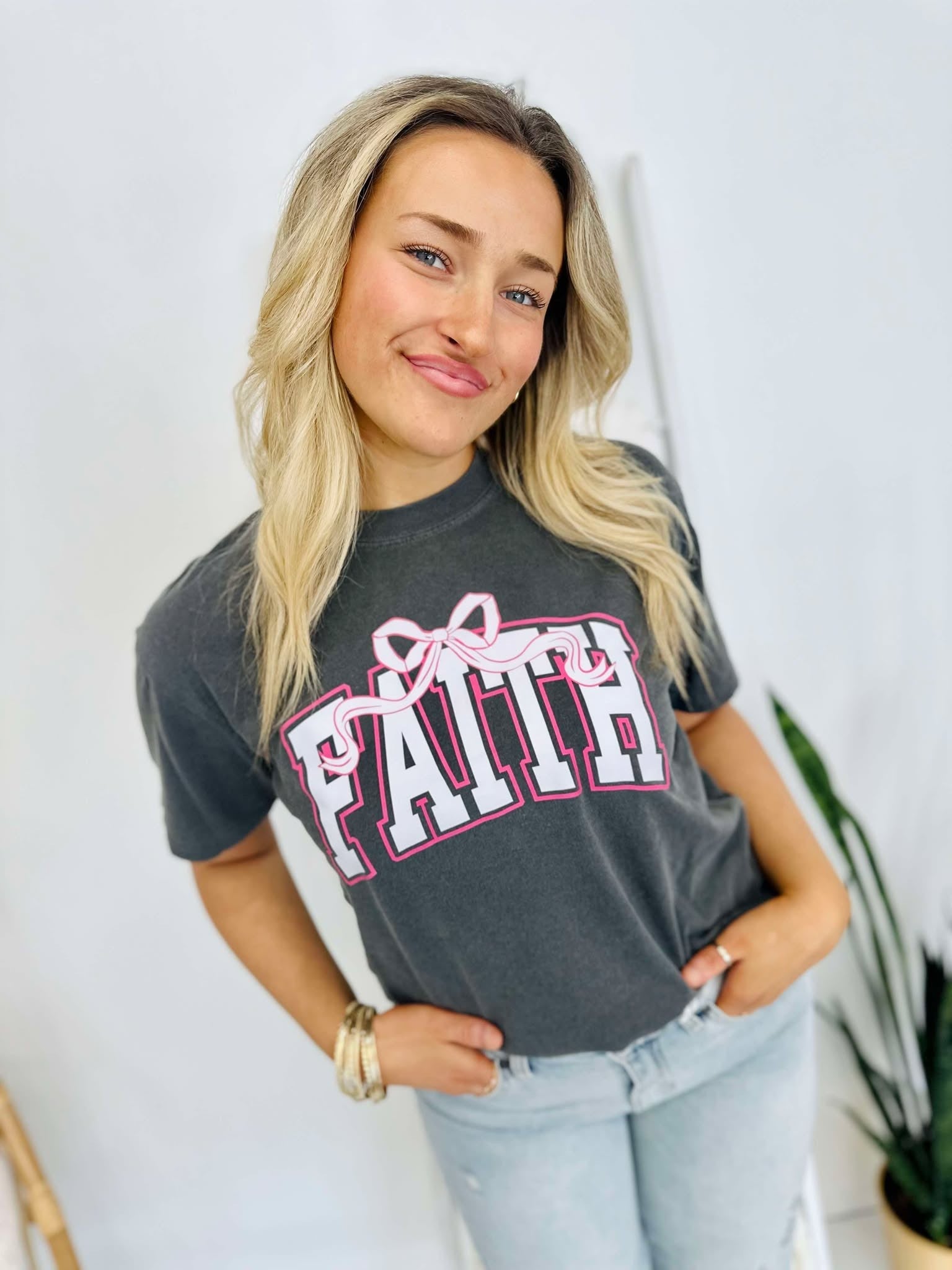 A woman wearing a gray t-shirt with the word 'Faith' and a pink bow on it, standing with one hand against a white background.