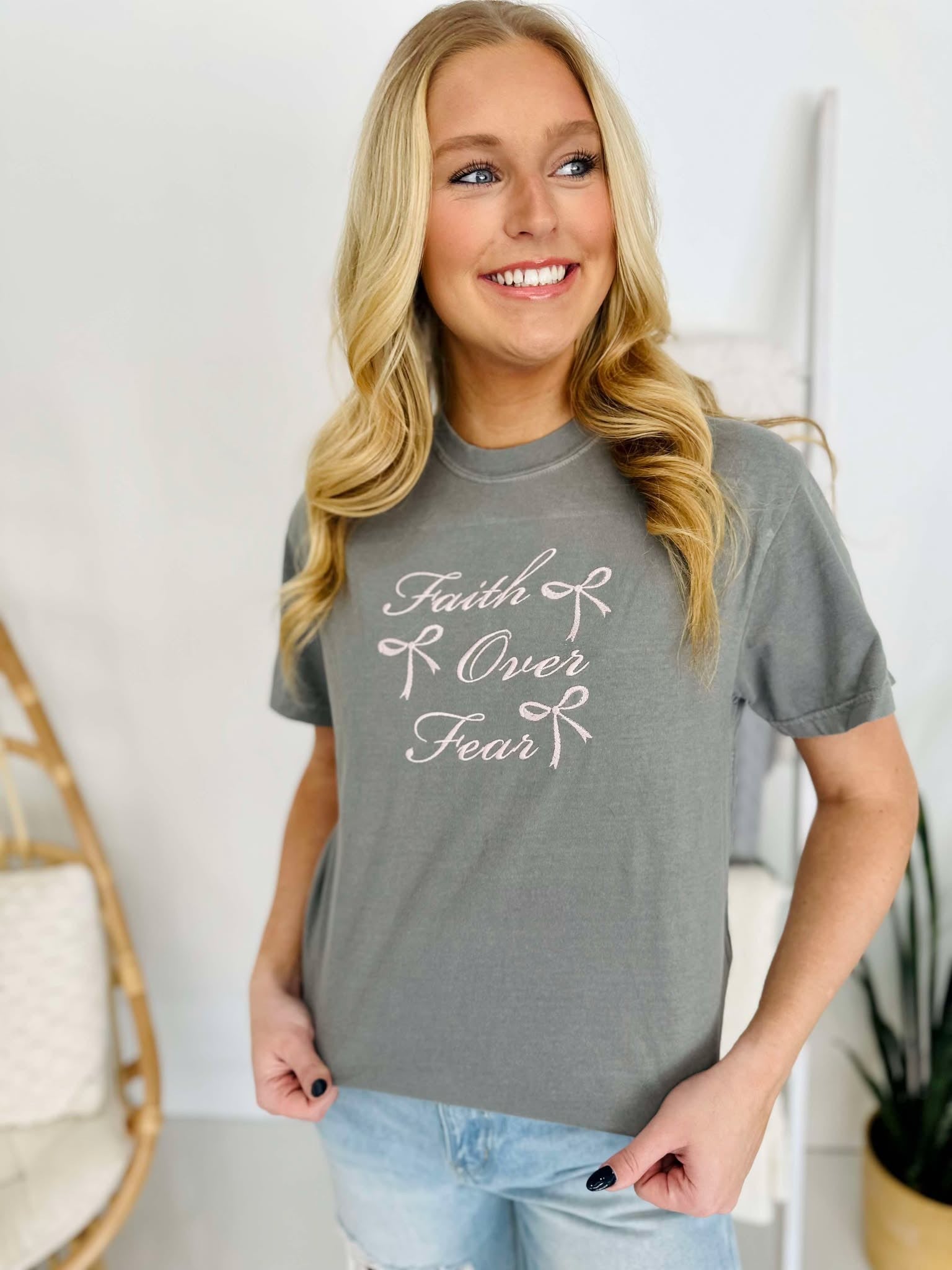 A woman smiling and wearing a gray t-shirt with 'Faith & Fear' embroidered on the front.