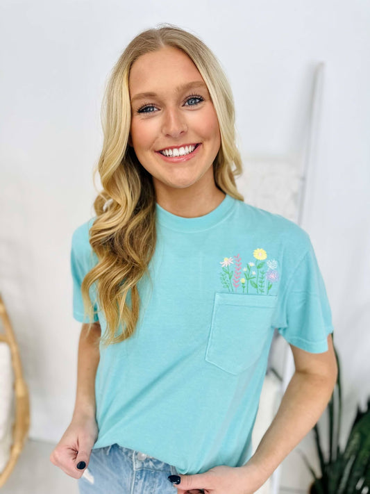 A woman wearing a chalky mint green t-shirt with a floral pocket design and embroidered lettering on the front, smiling against a white background.