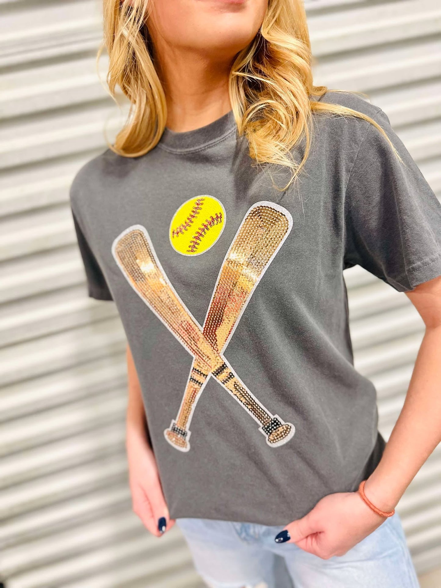 A female model wearing a gray t-shirt with a sequin patch on the front showing two softball bats and a yellow softball.