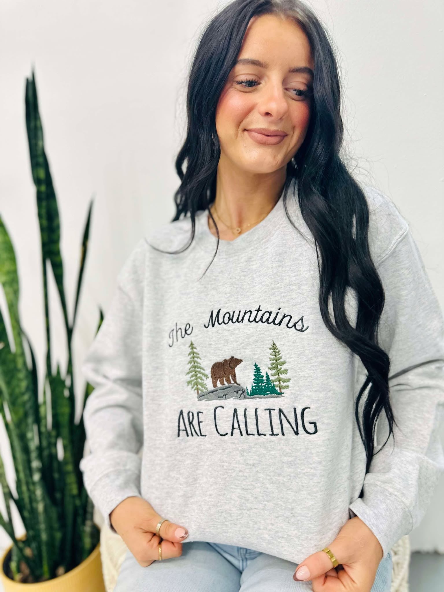 A woman smiling, wearing an ash grey embroidered sweatshirt with the text 'THE MOUNTAINS ARE CALLING' and an image of a bear in a forest.