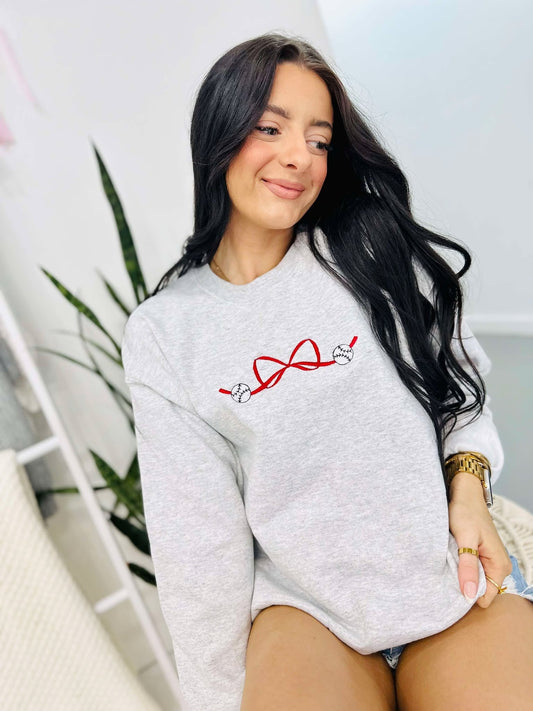 A woman smiling and sitting down, wearing a grey sweatshirt with a red and white embroidered bow design and the baseballs.