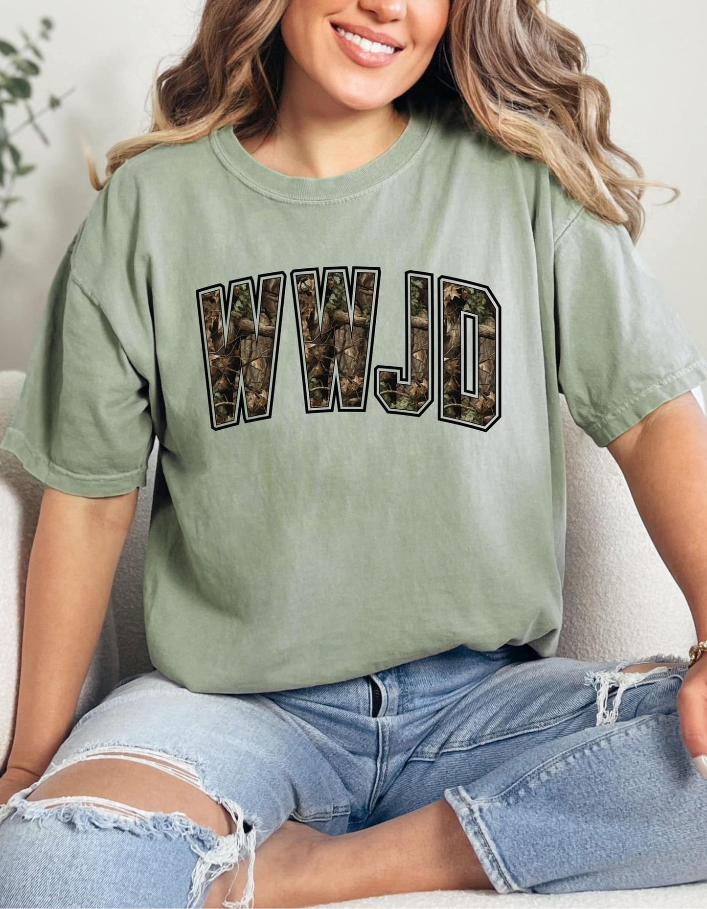 A person wearing a green t-shirt with 'WWJD' camo graphic text on it, sitting on a surface with blue jeans.