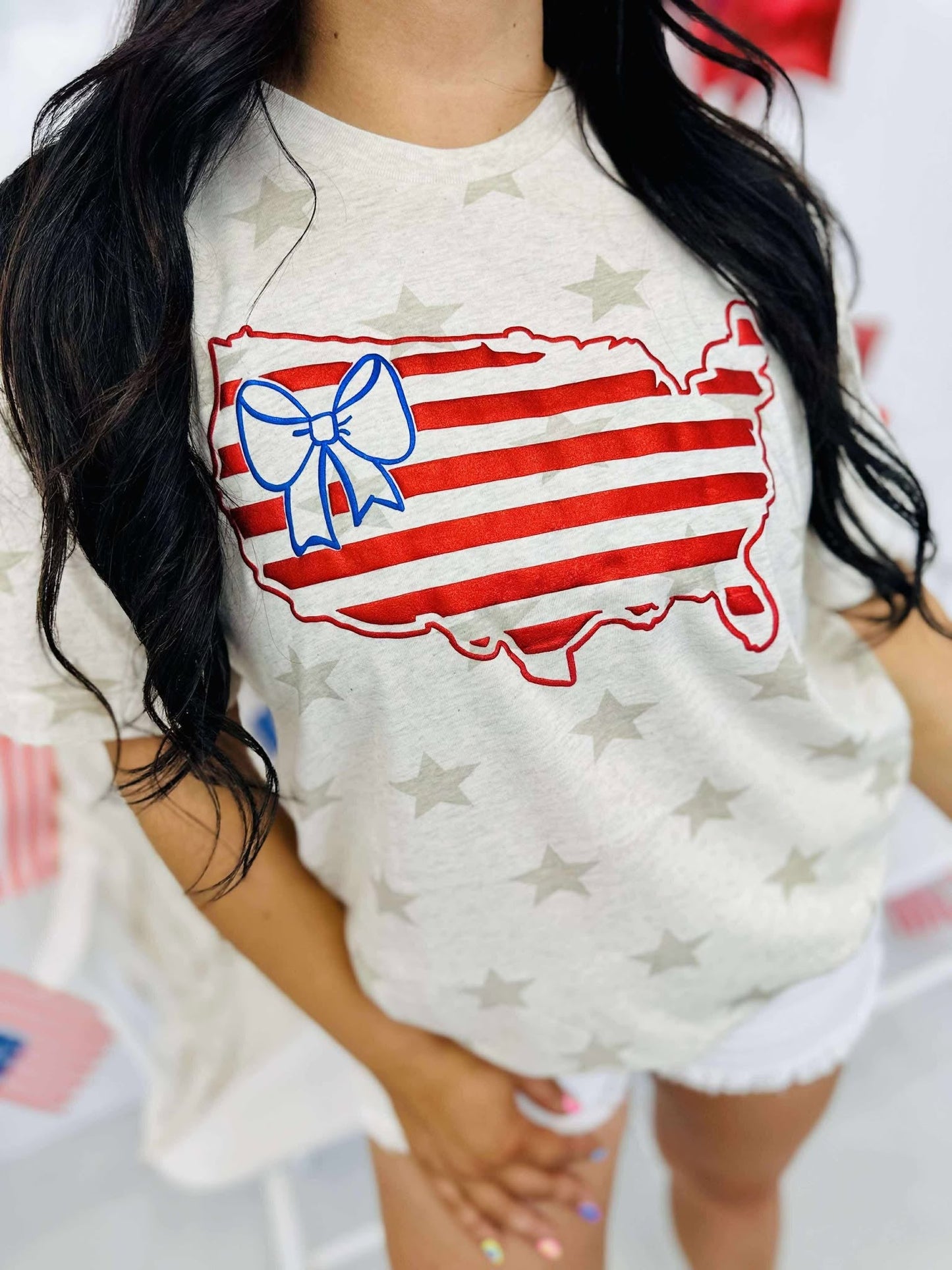 A graphic tee with a patriotic theme featuring an outline of the United States in red and blue colors with a bow on it, against a natural heather star pattern.