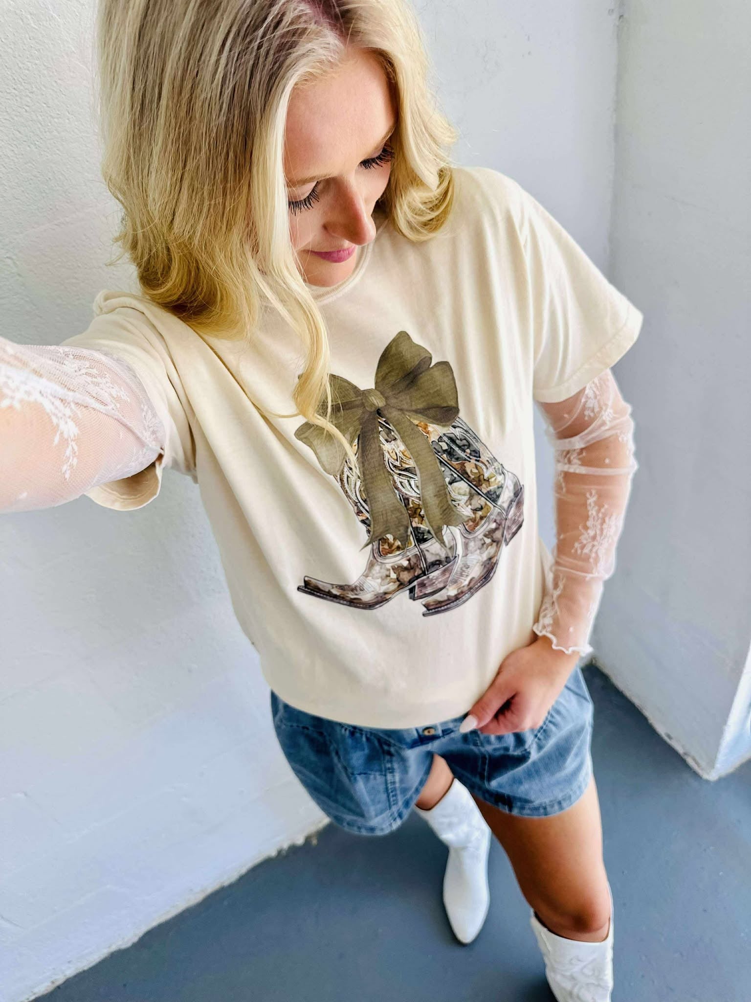 A graphic tee with a camo bow and boots design, predominantly in beige color, paired with blue denim shorts and white boots.