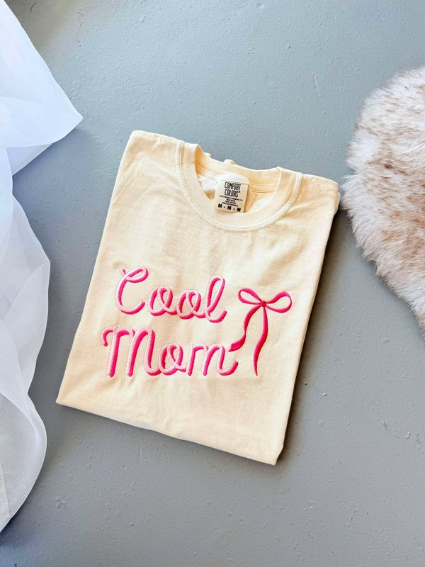 An ivory colored graphic tee with 'Cool Mom' embroidered in pink lettering, accompanied by a pink bow illustration.