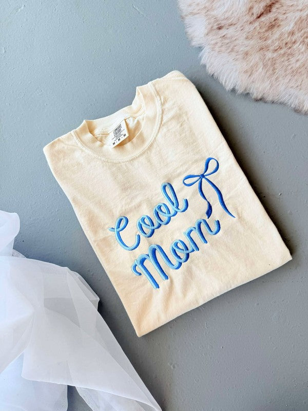 A yellow graphic tee shirt with blue embroidered lettering that reads 'Cool Mom' with a bow detail above the text.