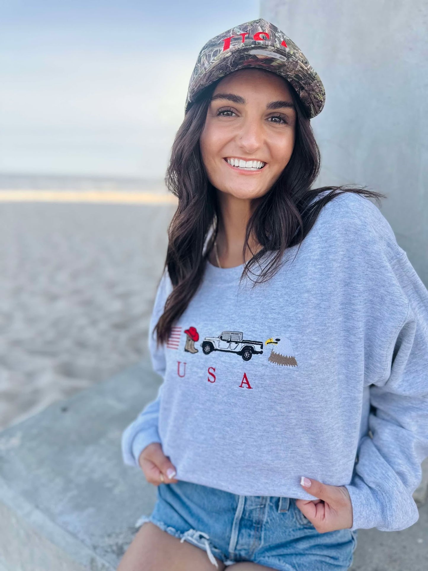 Woman wearing a light blue sweatshirt with an eagle, cowboy boots, truck and usa flag graphic design, sitting on a beach.